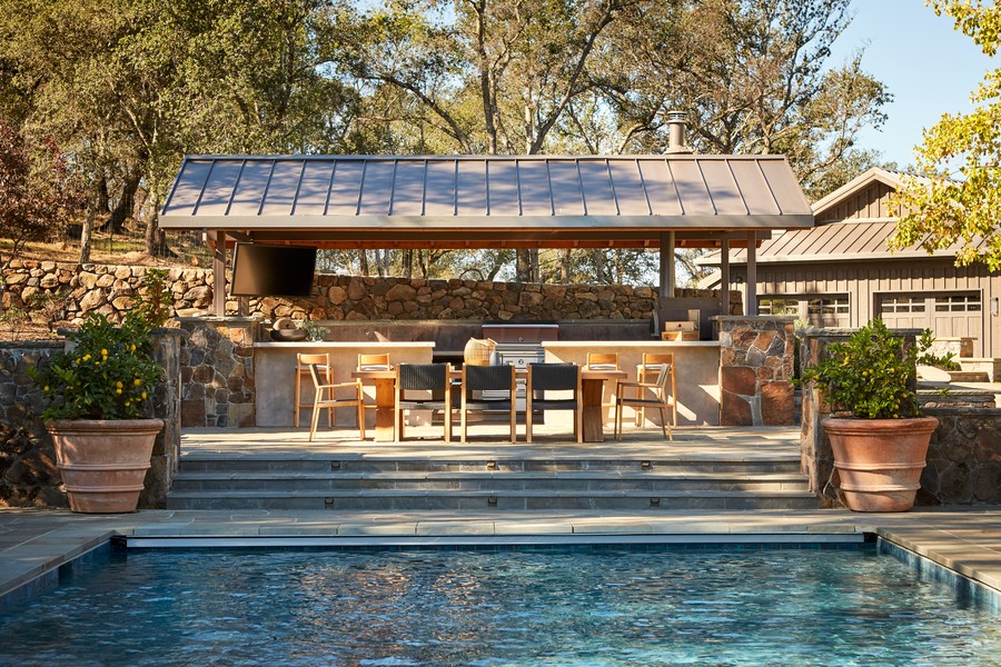 A roofed outdoor space near a pool, with a kitchen and outdoor TV.