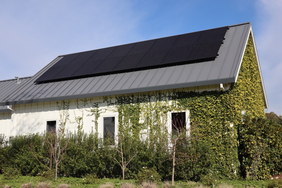 The image shows a large white building with a slanted roof covered in solar panels. The building is surrounded by dense greenery, including trees and vines growing up the walls.