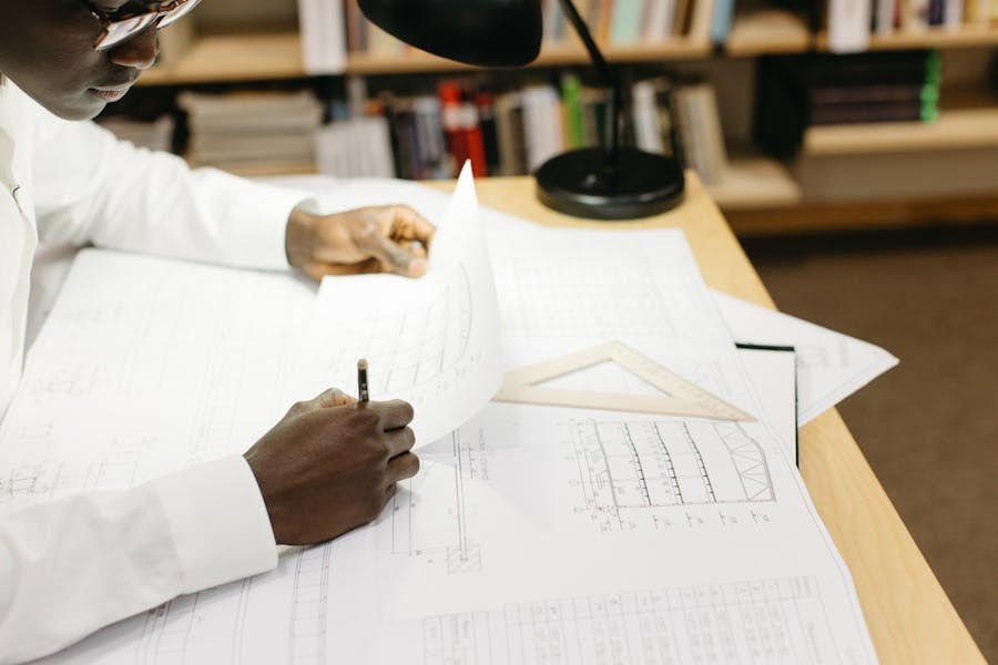 Architect working on detailed blueprints at a desk, surrounded by drafting tools and illuminated by a desk lamp, with bookshelves in the background.