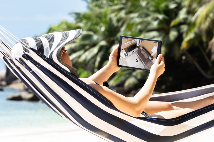 A person relaxing on a striped hammock at the beach, wearing a sun hat, and monitoring home security footage on a tablet, surrounded by tropical palm trees.