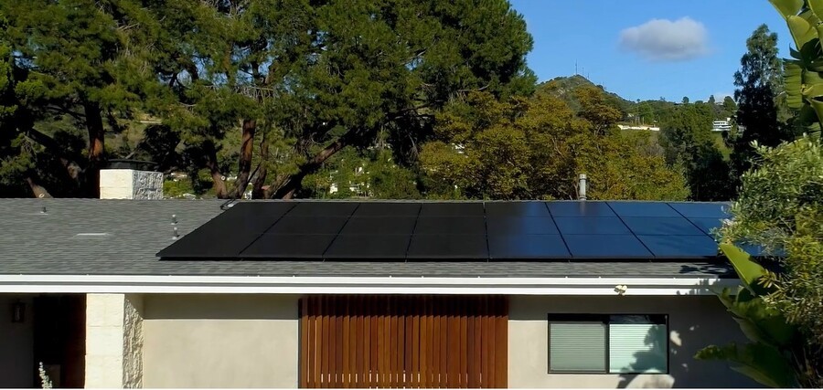 A modern single-story home features a sleek array of black solar panels installed on the roof, surrounded by tall trees and hills in the background.
