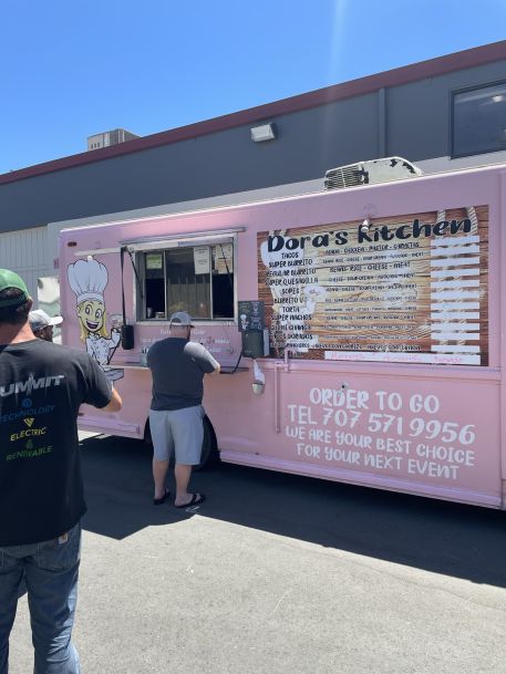 Pink food truck named “Dora’s Kitchen” parked in a lot on a sunny day, with two people standing at the service window reading the menu on the side of the truck.Pink food truck named “Dora’s Kitchen” parked in a lot on a sunny day, with two people standing at the service window reading the menu on the side of the truck.