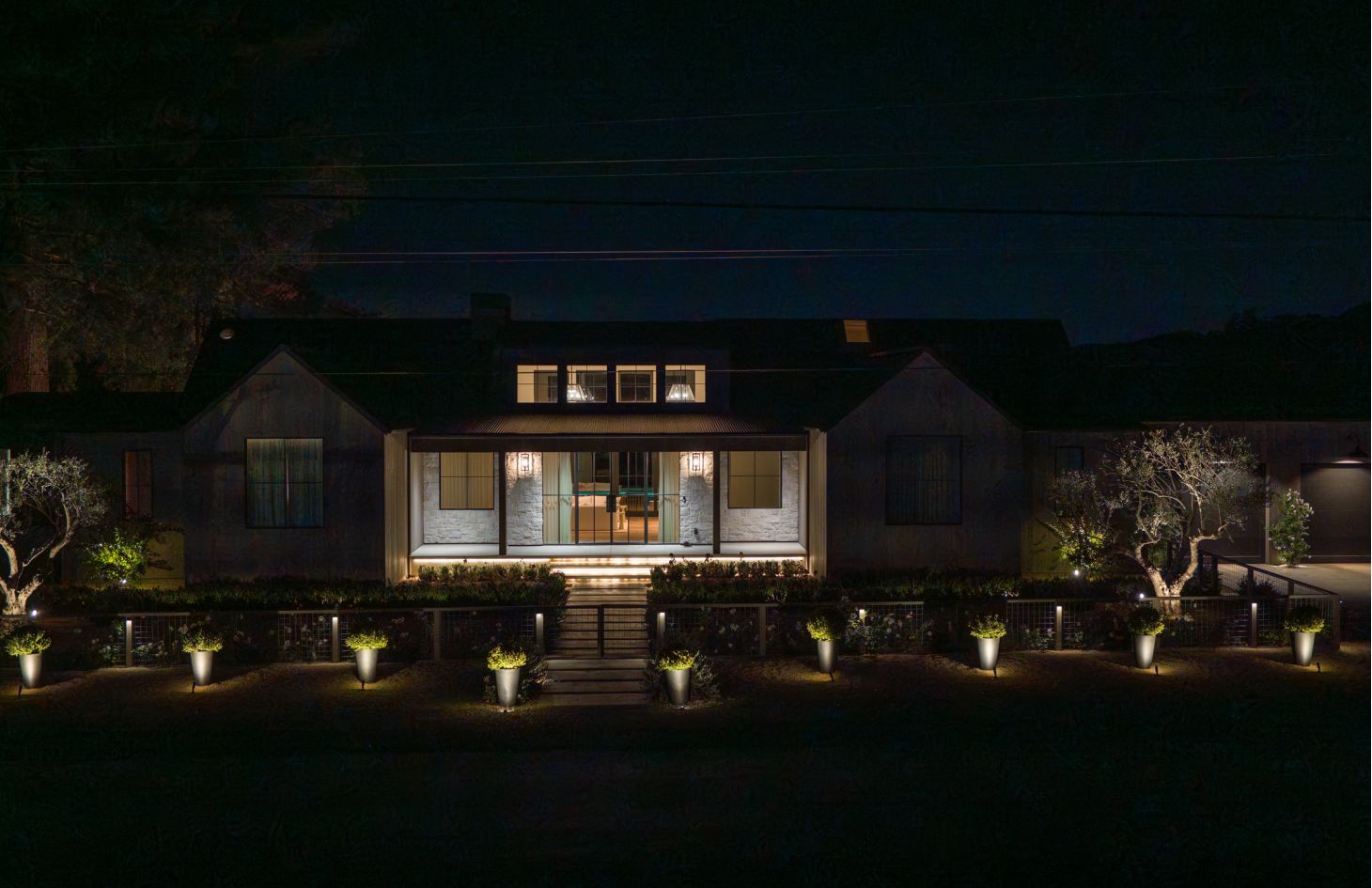 Front facade of the modern farmhouse at night, porch and pathway lights casting warm glows on stone columns, planters, and trees.