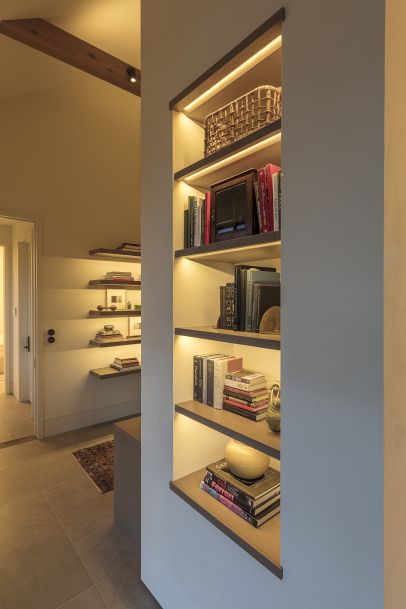 Built-in wall shelves with warm accent lighting displaying books and decorative objects in a modern hallway, with additional floating shelves and a rug visible in the adjoining room.