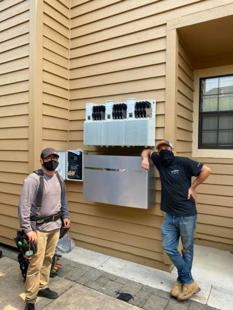 Two technicians standing next to a wall-mounted electrical system with multiple inverter units installed on the exterior of a home.