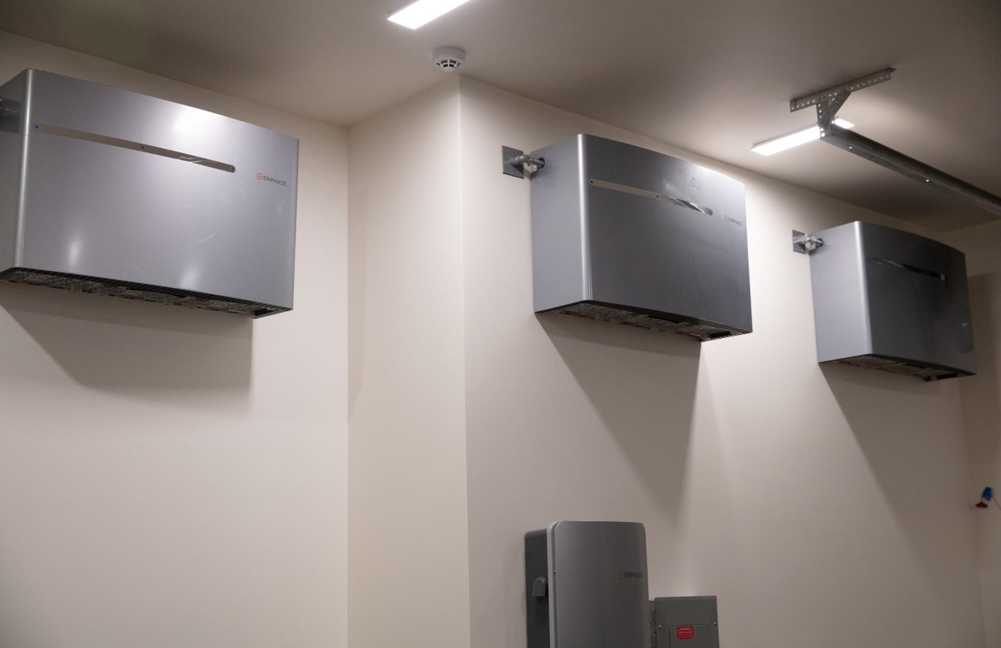Three wall-mounted gray battery backup units lined up high on a clean garage wall under slender LED ceiling lights.