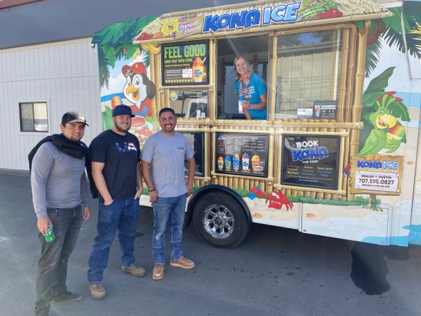 Three men stand smiling in front of a colorful Kona Ice shaved ice truck, with a server leaning out of the service window and tropical graphics decorating the side.