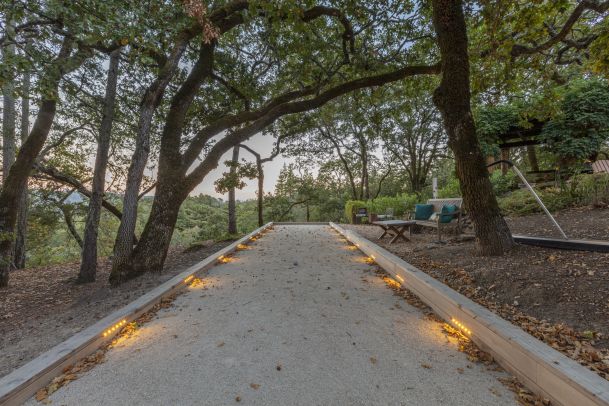 A gravel walkway lined with integrated landscape lighting, surrounded by trees and natural scenery, leading to an outdoor seating area.