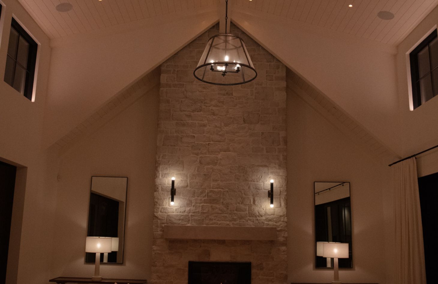 Warm, low-level lighting highlights a vaulted-ceiling great room with a floor-to-ceiling stone fireplace, twin wall sconces, modern console tables with white drum-shade lamps, and tall mirrors flanking the hearth.