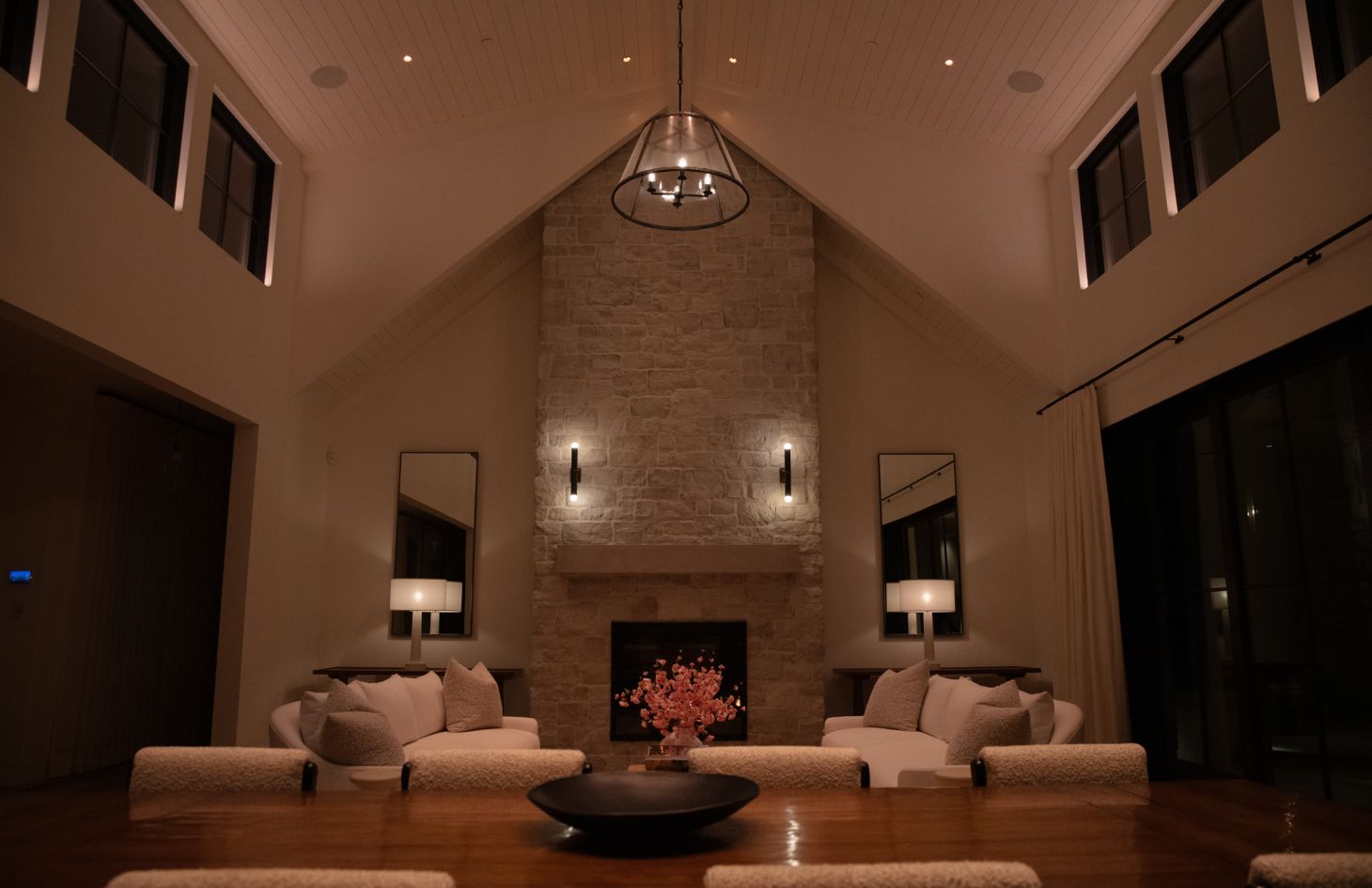 Symmetrical frontal view of a vaulted living room: stone fireplace centered between tall mirrors and table lamps, twin white sofas with textured pillows, and dim uplighting that frames high windows and ceiling beams.