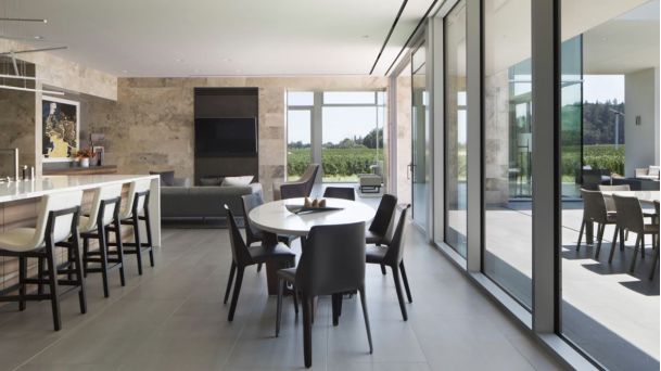 A modern open-concept kitchen and dining area with a large island, bar seating, and floor-to-ceiling sliding glass doors overlooking an outdoor patio and scenic view.