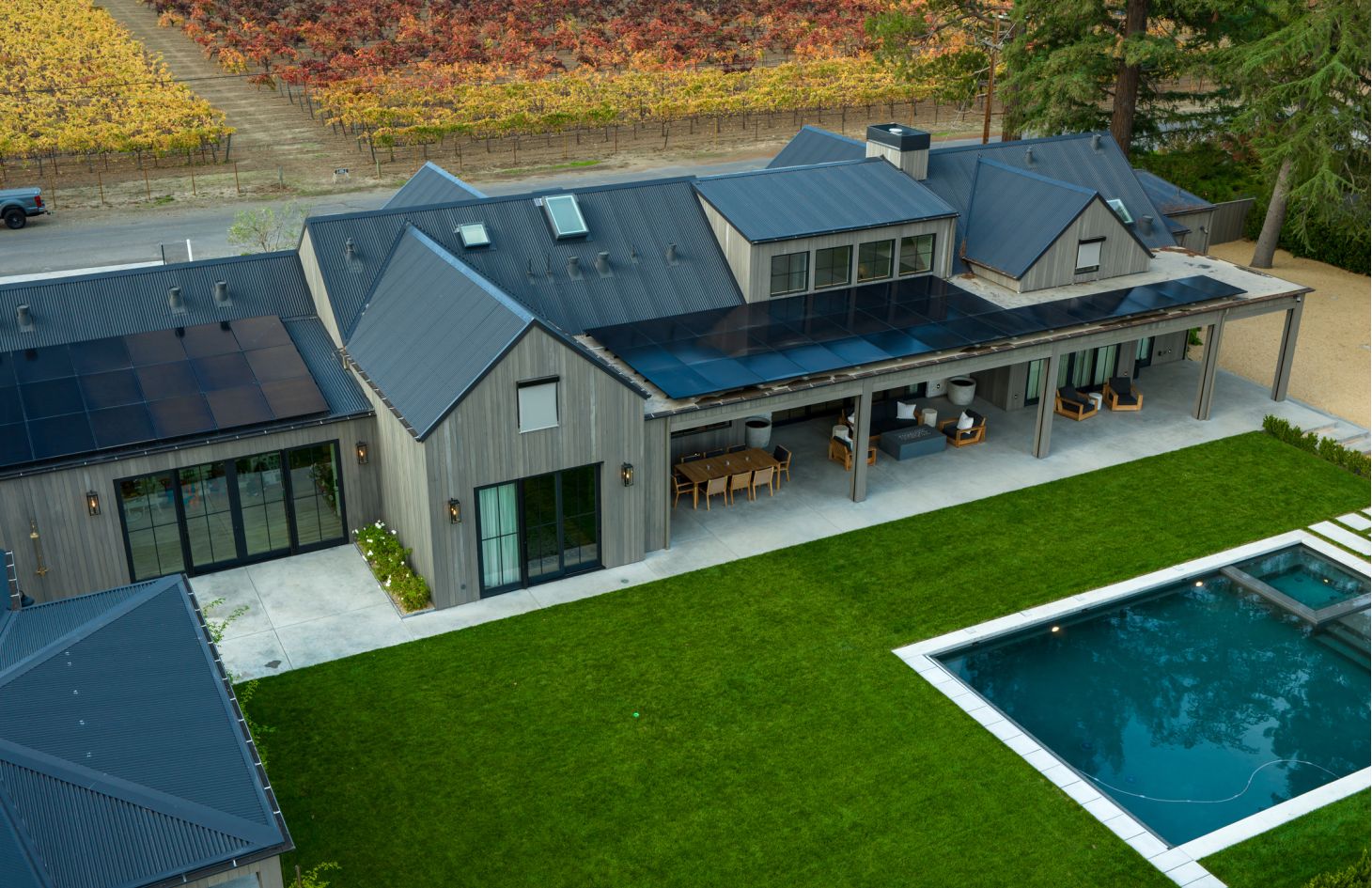 Oblique aerial of the backyard: cedar-clad house with solar-panel patio cover, outdoor dining set, lush lawn, and pool beside colorful autumn vineyards.