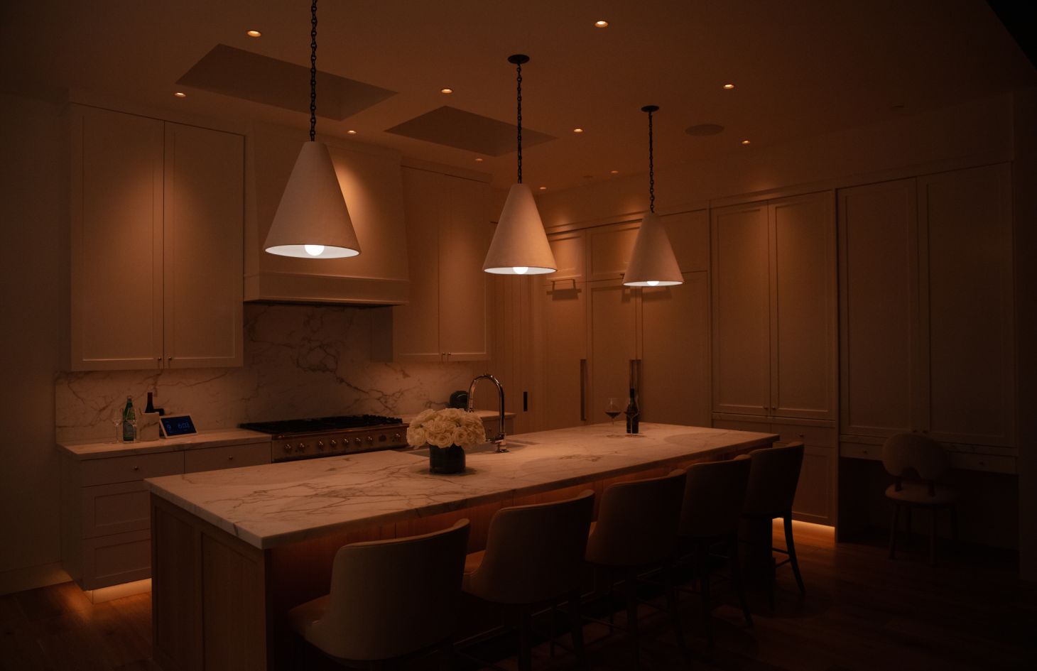 Moody evening scene of the marble-island kitchen: three cone pendants glow softly over the counter while toe-kick lights and recessed cans provide subtle accent lighting.