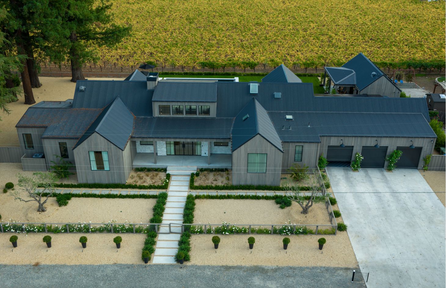 Elevated daytime shot of the home’s front: gray wood siding, three-bay garage, wide gravel beds, clipped hedges, and a centered stepping-stone walkway.